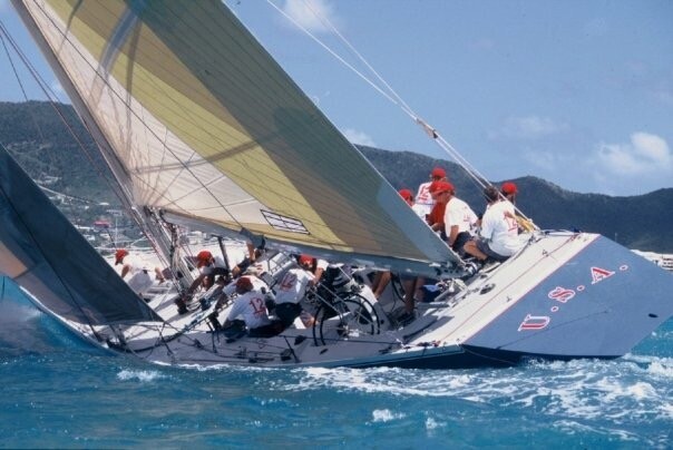A 12 Metre yacht heeled hard over under full crew during a race, sails yellow and white against blue water, hillside behind